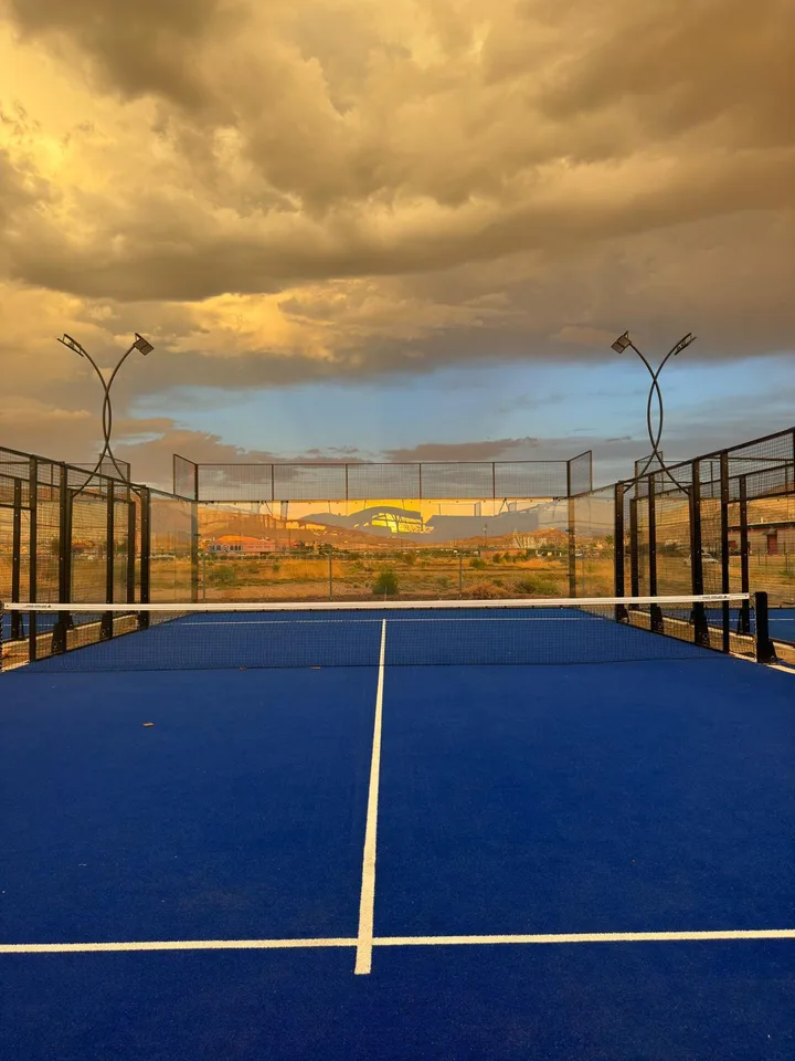 Blue padel court beneath a golden sunset, with glass walls and curved light posts.