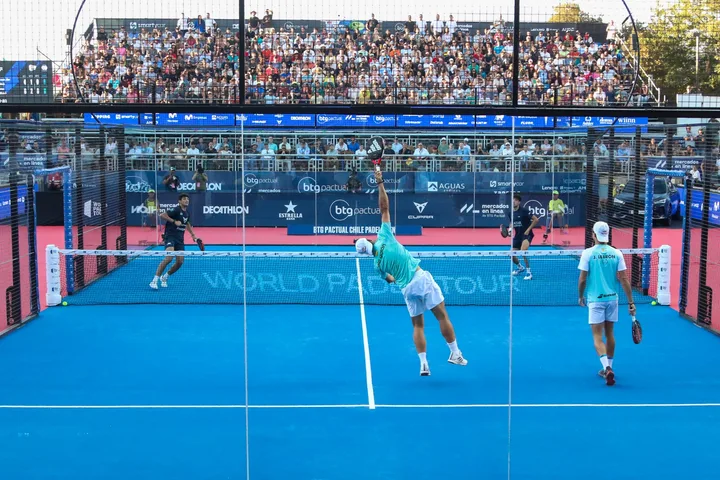 Four players during a padel match on a blue court with spectators in the stands.