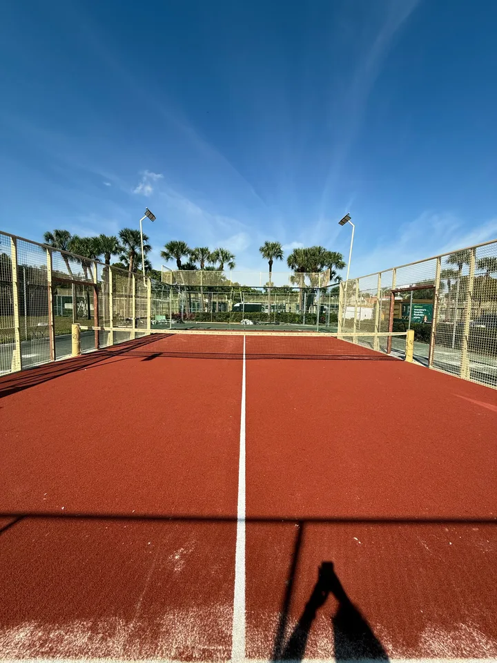 Terracotta-colored padel court with pale mesh fencing and palm trees behind it.